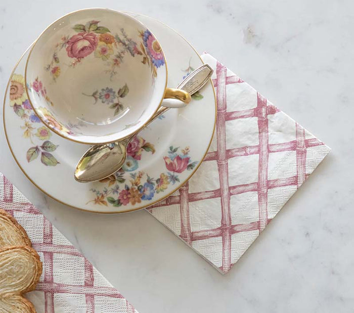 A floral teacup with saucer and silver spoon sits on Hester & Cook Pink Lattice Cocktail Napkins (pack of 20) beside slices of bread, creating an inviting cocktail hour scene on a white marble surface.