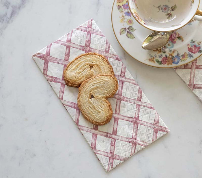 Two heart-shaped puff pastries rest on a Hester & Cook Pink Lattice Guest Napkin (pack of 16) beside a floral teacup and saucer on a white marble surface.