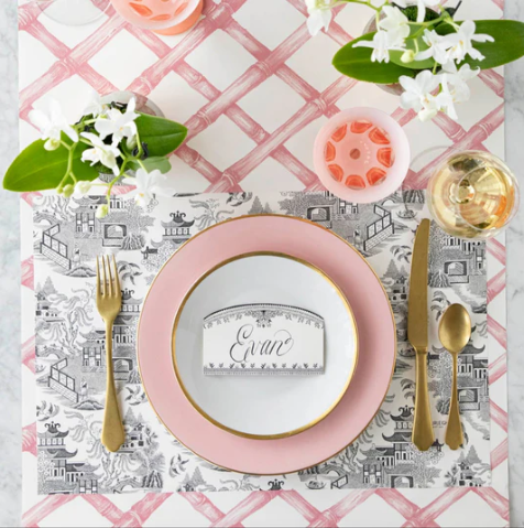 A chic table setting features gold cutlery, a pink and white plate, a place card for Evan, a wine glass, flowers in white vases, and a patterned placemat on the Hester & Cook Pink Lattice Runner (20x25").