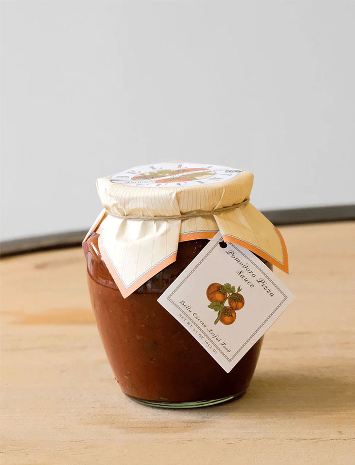 A glass jar of tomato sauce with a cream paper cover and tomato label sits on light wood—an ideal match for the Maple Personalized Pizza Peel Gift Set.