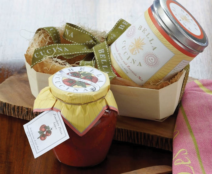The Personalized Pizza Peel Gift Set by Maple features a jar of tomato sauce and canister, stylishly labeled, and is adorned with raffia, green ribbon, and a pink napkin on a wooden surface.