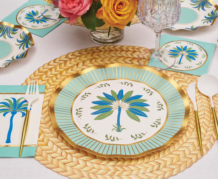 A festive table features Sophistiplate's Preppy Palm Dinner Plates (8pk) with a blue and green palm design, matching napkins and plates, gold cutlery, a woven placemat, and a vase of yellow and pink roses.