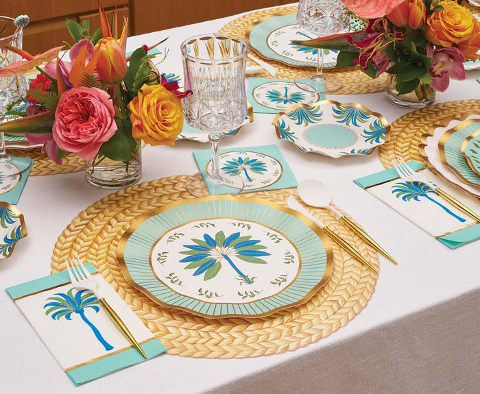 A table set with floral centerpieces, gold cutlery, crystal glasses, woven placemats, and napkins features Sophistiplate Preppy Palm Dinner Plates/8pk with a blue and gold design, all arranged on a white tablecloth.