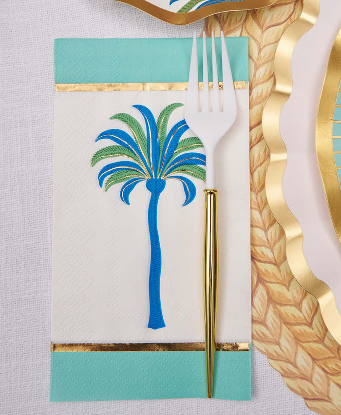 The Sophistiplate Preppy Palm Guest Towels/20pk, featuring a blue and green palm tree design with a gold border, are displayed on a teal-bordered napkin beside gold-accented plates and a white and gold fork on a textured surface.