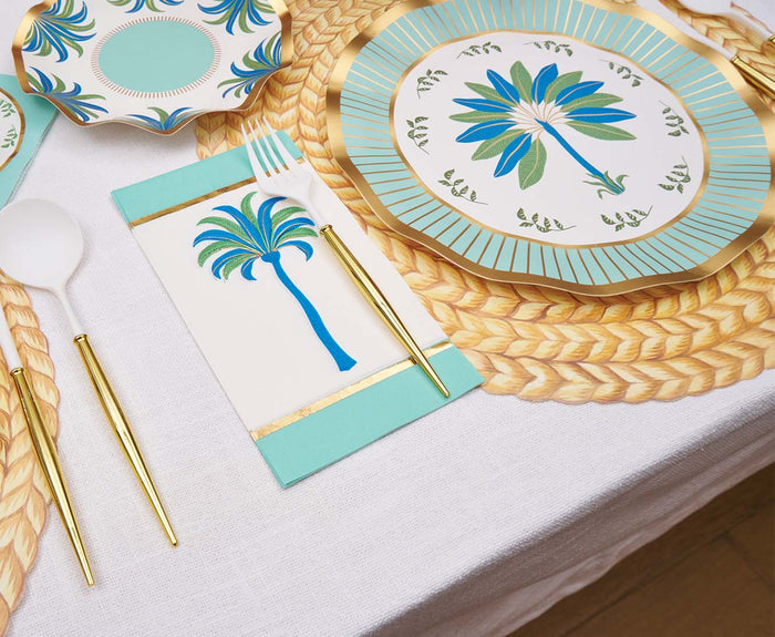 A table setting with a white tablecloth, gold and white cutlery, blue and green palm leaf plates, a woven placemat, and Sophistiplate Preppy Palm Guest Towels (20pk) featuring a blue palm tree.
