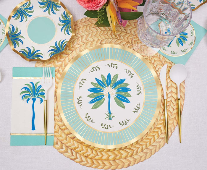 A table set with tropical plates, palm tree designs, gold utensils, a clear glass, woven placemat, and a floral centerpiece on a white tablecloth is accented by Sophistiplate Preppy Palm Guest Towels (20pk) for a chic summer vibe.