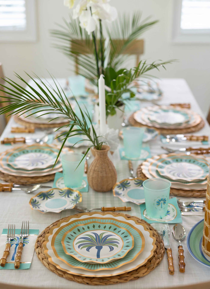 The Preppy Palm Table Setting by Sophistiplate LLC features tropical-themed plates, bamboo cutlery, woven placemats, blue cups, palm leaf accents, and a white orchid vase—perfect for a fresh, summery table.