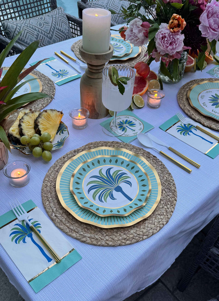 A close-up of the Preppy Palm Table Setting by Sophistiplate LLC features palm-themed plates, gold cutlery, a candle, floral centerpiece, tropical fruits, and teal napkins on woven placemats and a white tablecloth.