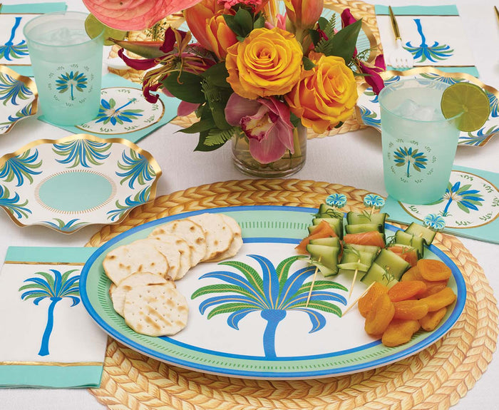 The Ultimate Preppy Palm Tablescape by Sophistiplate LLC showcases designer palm tree paper plates, cups, and napkins. A platter holds crackers, dried apricots, and cucumber beside a vase of orange and yellow flowers at the center.
