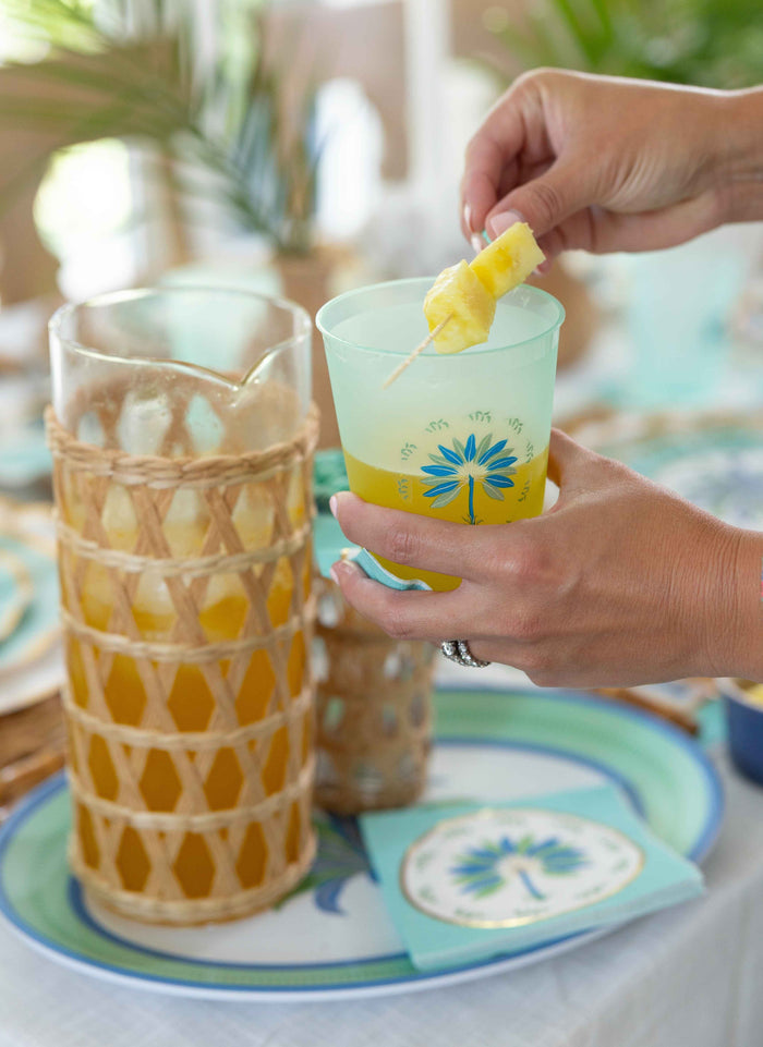 A person holds a green floral glass with a yellow drink and pineapple garnish; the Sophistiplate LLC Preppy Palm Table Setting, including a matching pitcher and sturdy plates, sits nearby—perfect for any party-in-a-box celebration.