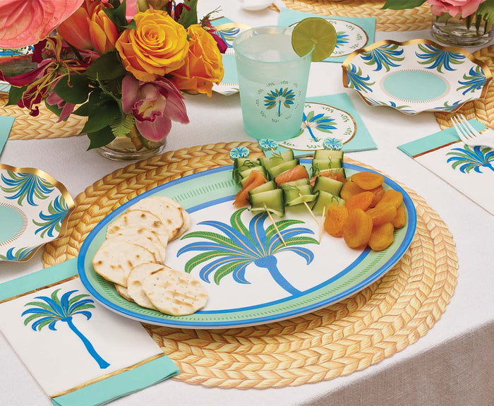 A tropical table is set with a Sophistiplate Preppy Palm Melamine Serving Platter holding crackers, cucumber skewers, and dried apricots, complemented by a floral centerpiece, matching napkins, and a cup garnished with lime.