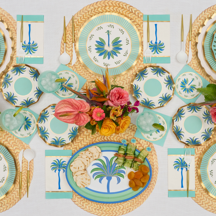 A vibrant table features palm-themed plates, a Sophistiplate Preppy Palm Melamine Serving Platter, gold utensils, woven placemats, teal glasses with ice, floral napkins, and a tropical flower centerpiece. Some plates hold flatbreads and skewers.