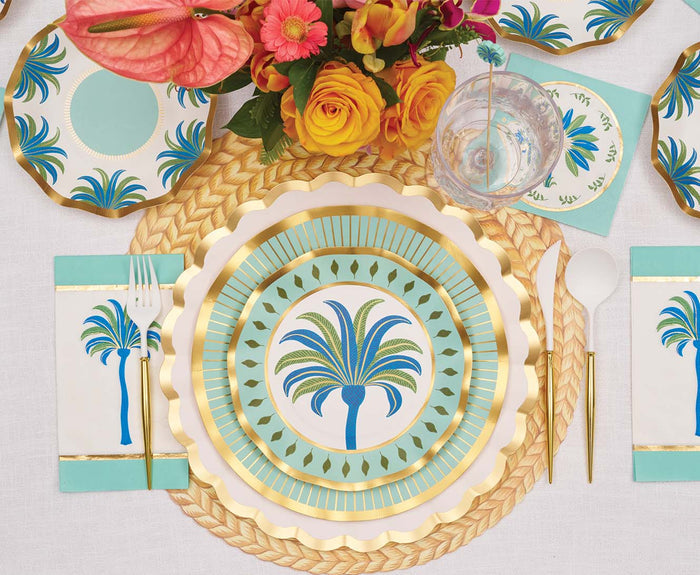 A table set with Sophistiplate Preppy Palm Salad Plates (8pk), a woven placemat, gold utensils, a glass with clear straw, teal napkins, and a pink and yellow floral centerpiece creates a chic tropical vibe.