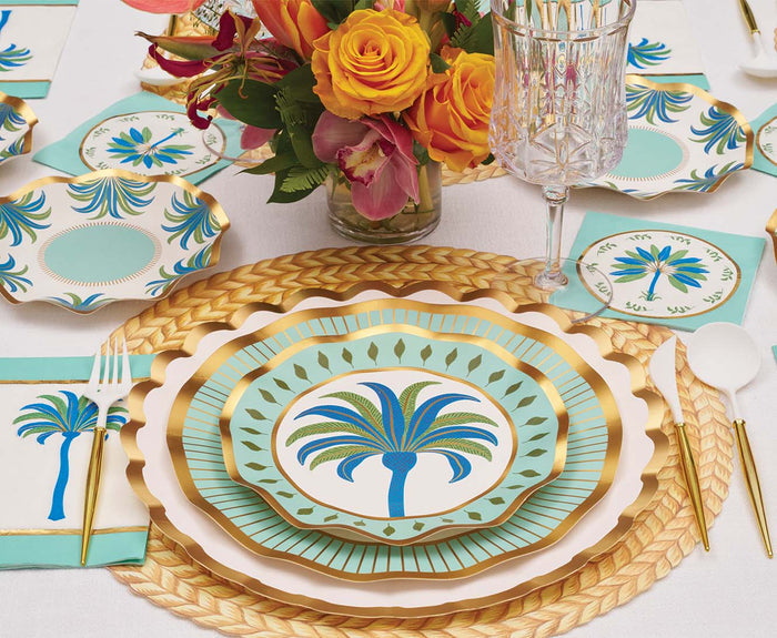 A table setting features Sophistiplate Preppy Palm Salad Plates (8pk) with palm tree designs, gold cutlery, a woven placemat, crystal glassware, floral napkins, and a vibrant orange and yellow flower centerpiece.