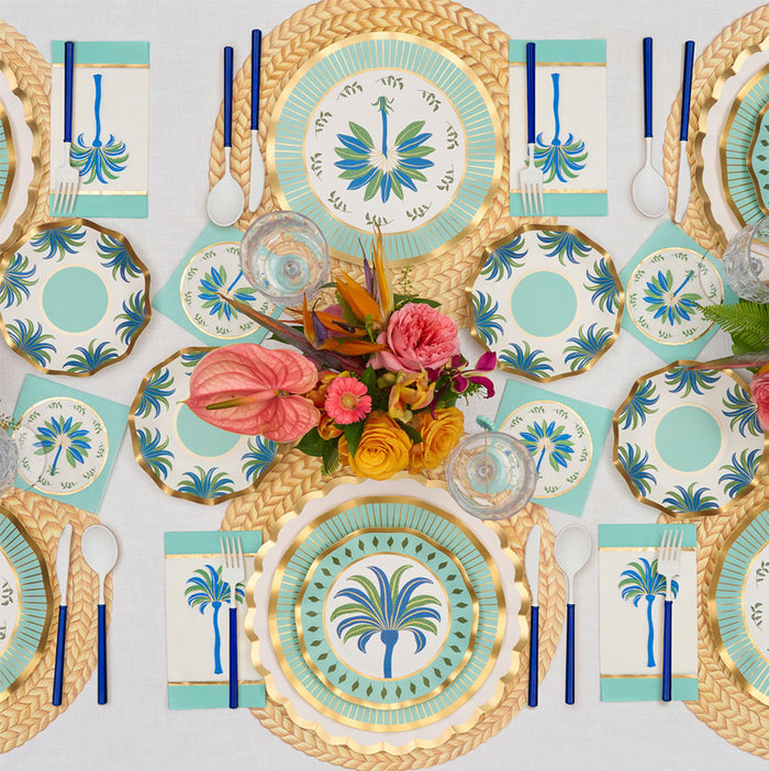 The Preppy Palm Table Setting by Sophistiplate LLC features gold placemats, palm tree-themed heavy-duty plates, blue-handled cutlery, and floral centerpieces with pink and orange blooms—all perfectly arranged on a white tablecloth.