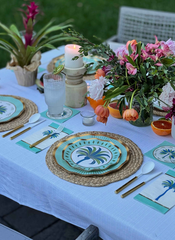 The Preppy Palm Table Setting by Sophistiplate LLC features tropical heavy-duty paper plates, gold cutlery, woven placemats, and more—all beautifully arranged on a white tablecloth for a stylish outdoor look.