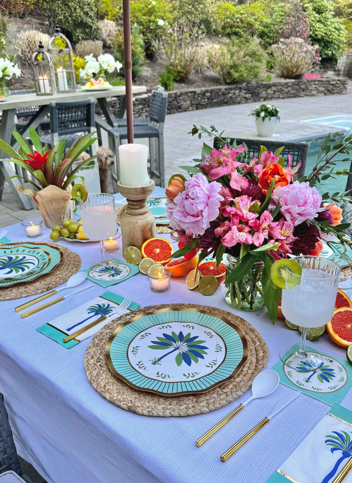 A vibrant outdoor table set with Sophistiplate LLC's Preppy Palm Table Setting—disposable party plates, gold cutlery, candles, drinks, citrus slices, and pink flowers on a white cloth—in a sunny garden.