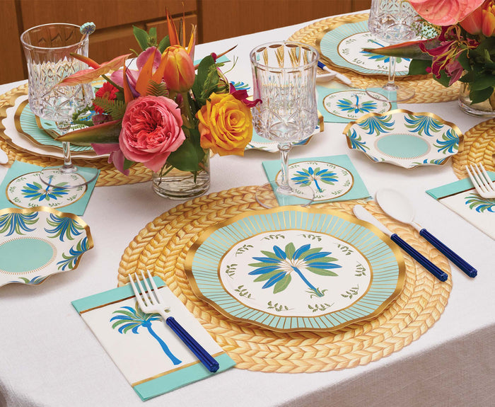 The Preppy Palm Table Setting by Sophistiplate LLC features festive disposable party plates, blue-handled cutlery, woven placemats, floral centerpieces, and palm tree napkins for the perfect tropical party-in-a-box celebration.