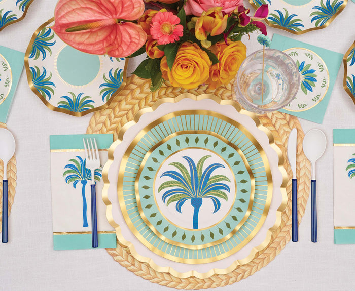 The Preppy Palm Table Setting by Sophistiplate LLC features heavy duty palm tree paper plates with gold rims, matching napkins, blue-handled cutlery, and bright pink, yellow, and orange flowers on woven placemats.