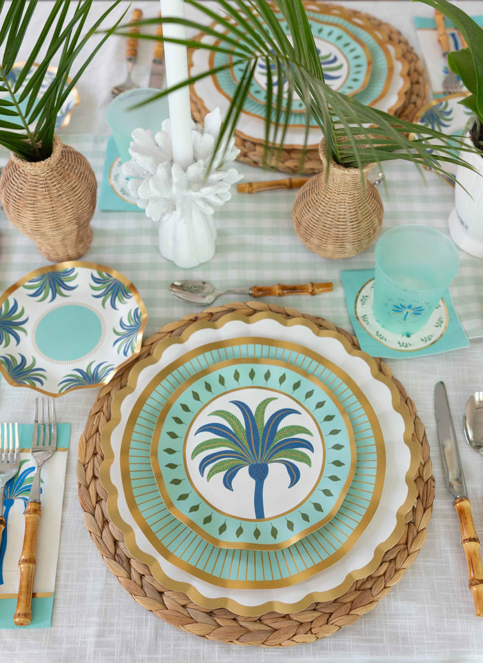 The Preppy Palm Table Setting by Sophistiplate LLC features woven chargers, palm tree paper plates, bamboo cutlery, pale blue glasses, and vases with palm leaves on a white patterned tablecloth for a tropical vibe.