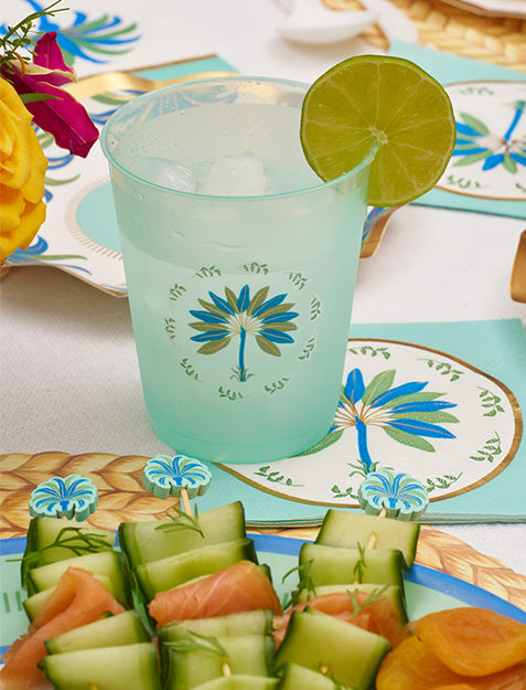 A light blue drink with ice and lime in a decorated cup featuring a blue and green palm motif is part of the Preppy Palm Table Setting by Sophistiplate LLC, complete with matching napkins, heavy duty paper plates, and cucumber-wrapped appetizers.