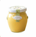 A glass jar of Bella Cucina Preserved Lemon Cream, a citrus spread with preserved lemons, is sealed with a white lid, decorated with a green ribbon and oval label, and displayed on a white background.