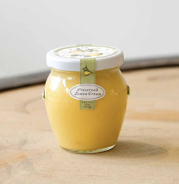A glass jar of Bella Cucina Preserved Lemon Cream sits on a wooden surface, topped with a white lid and a label featuring an illustrated lemon.