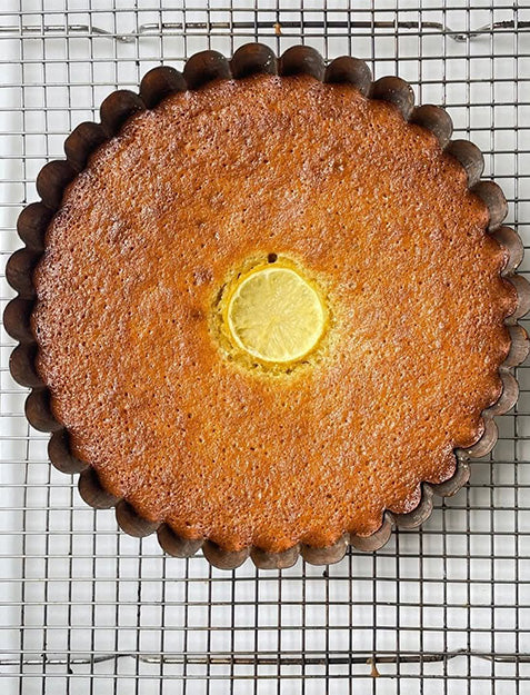 A round, golden-brown cake with scalloped edges sits on a cooling rack, topped with a single lemon slice and a touch of Bella Cucina Preserved Lemon Cream citrus spread in the center.