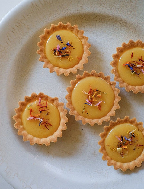 Five small tartlets with golden yellow Bella Cucina Preserved Lemon Cream filling and scalloped edges, topped with colorful dried flower petals, are arranged on a white, textured plate.