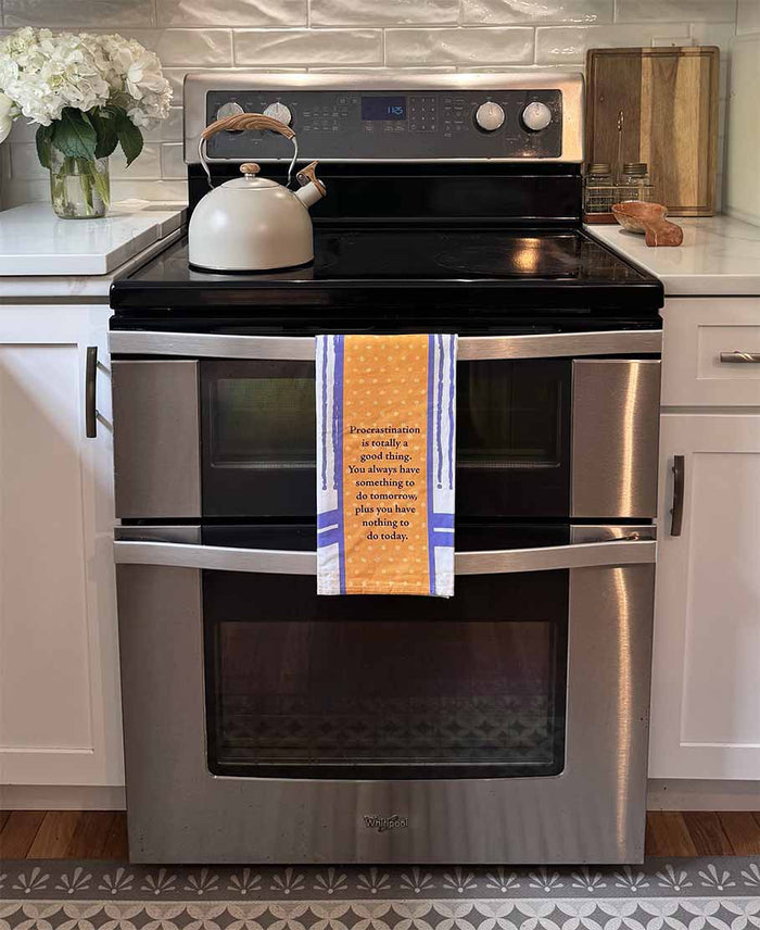 A modern kitchen features the Drinks On Me Procrastinations Towel, a tea kettle on the stove, stainless steel oven, white cabinets, cutting boards, a flower vase, and white tile backsplash—perfect as a housewarming gift.