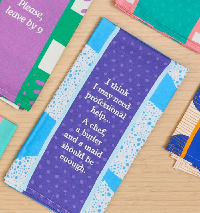 The Professional Help Dish Towel by Drinks On Me features blue and purple tones with fun white text. Placed on a light wood surface, it makes a perfect humorous kitchen gift alongside other colorful dish towels.