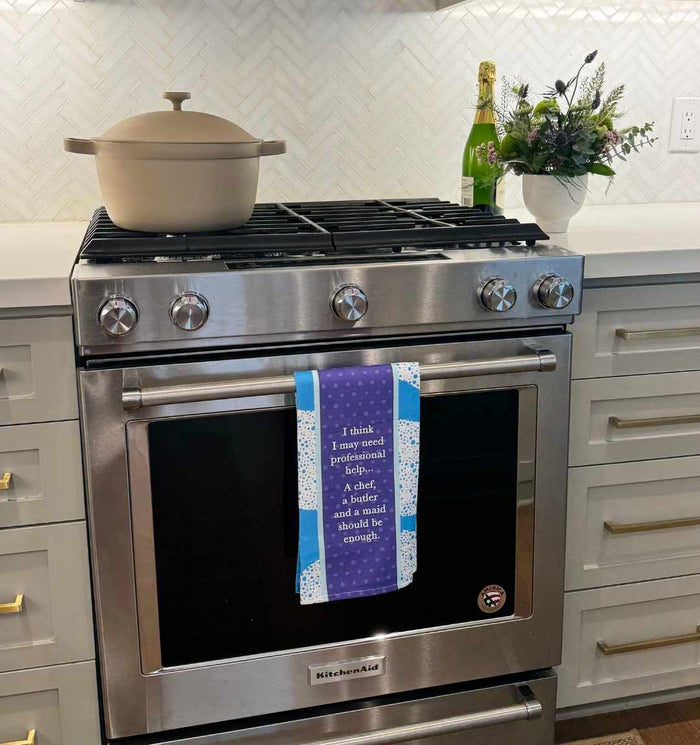 A modern stainless steel oven displays the Drinks On Me Professional Help Dish Towel with a witty cooking quote, making it a fun kitchen gift. Nearby are a beige Dutch oven, sparkling wine, and flowers on the counter.