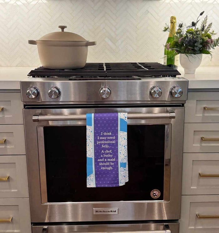 A modern stainless steel oven with a covered pot on the stove, the Professional Help Dish Towel by Drinks On Me hanging from the oven handle, and a champagne bottle with flowers on the counter. White cabinets and a herringbone backsplash complete the kitchen.