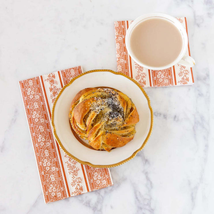 A cup of coffee with milk and a poppy seed pastry sit on Hester & Cook Pumpkin Burleigh Stripe Cocktail Napkins, adding a festive touch to the white marble surface.
