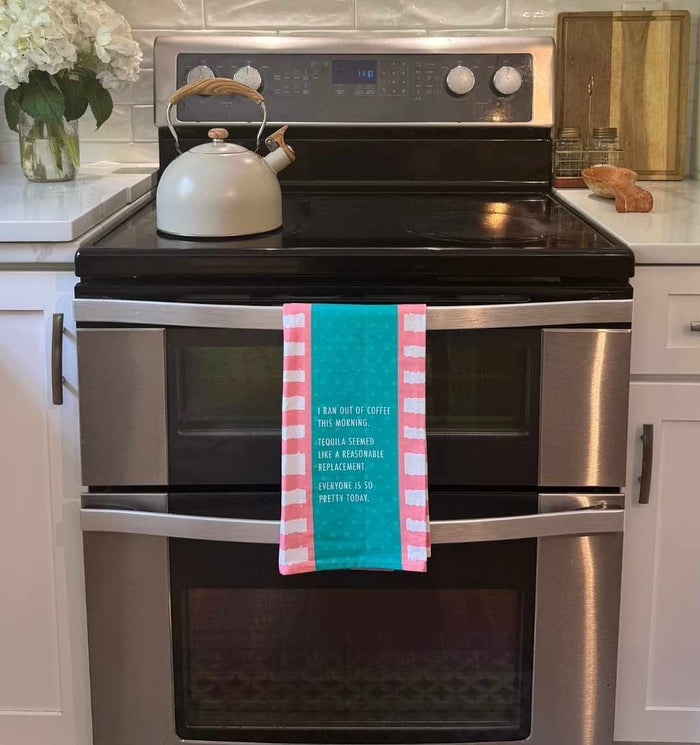 The Drinks On Me "Ran Out of Coffee" dish towel—striped in teal and pink—hangs on a stainless steel oven, adding quirky charm to a white kitchen with a teapot and flowers. Perfect for kitchen humor or as a fun housewarming gift!.