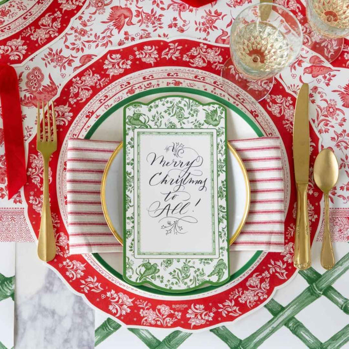A festive Christmas table setting with gold cutlery, a red and white plate, striped napkin, crystal glasses, Hester & Cook Red Burleigh Placemats (12ct), and a “Merry Christmas to All!” card on a green and white patterned plate.