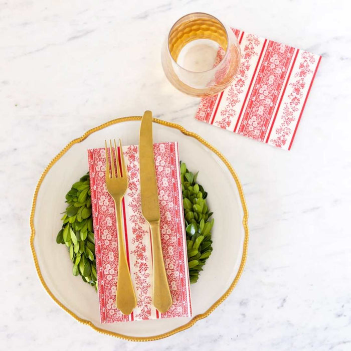 A table is set with a gold-rimmed plate, gold flatware, textured glass, a green wreath, and Hester & Cook Red Burleigh Stripe Cocktail Napkins (20pk), all elegantly displayed on a white surface.