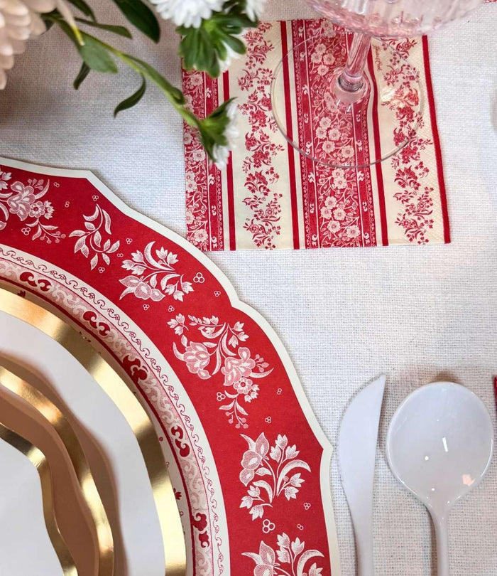 A close-up of the Red Burleigh Table Setting by Sophistiplate features red and white floral-patterned disposable plates, a matching napkin, gold-rimmed plate, cup, white cutlery, and a pink glass on a flower-adorned white tablecloth.