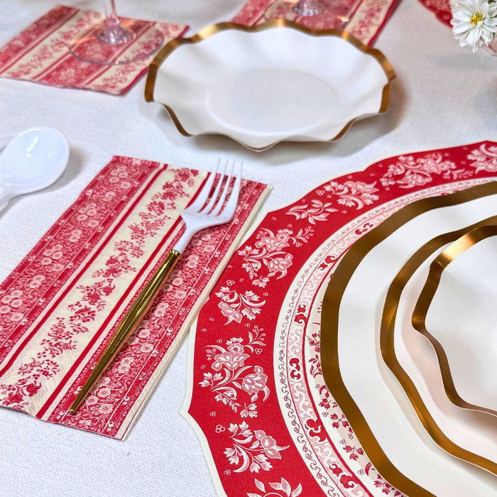 The Red Burleigh Table Setting by Sophistiplate features gold-edged white disposable plates, red and cream floral placemats, gold-accented cutlery, and a white plastic spoon for an elegant all-in-one tablescape.