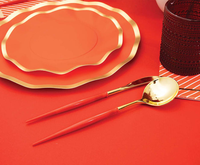 Sophistiplate’s Red & Gold Bella Plastic Cutlery (24pc, service for 8) is arranged beside stacked red and gold plates on a red table, with a dark textured glass and red-and-white striped napkin accenting the festive scene.