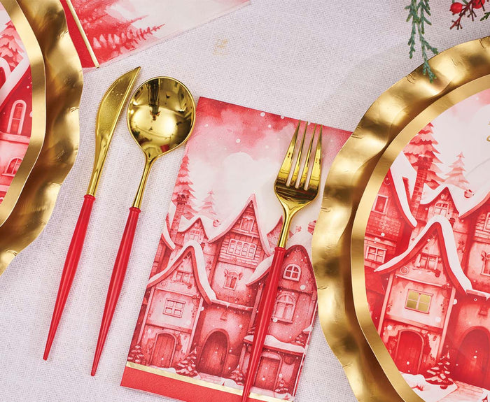 A festive table is set with Sophistiplate's Red & Gold Bella Plastic Cutlery (24pc, Service for 8), gold-rimmed plates, and a napkin with red illustrated snow houses, all on a white tablecloth.