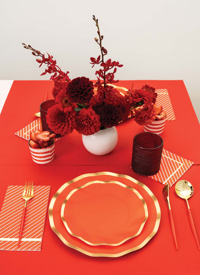 A table setting featuring Sophistiplate's Red & Gold Bella Plastic Cutlery (24pc, Service for 8), red and gold plates, striped napkins, red glassware, and a white vase with red flowers on a red tablecloth.