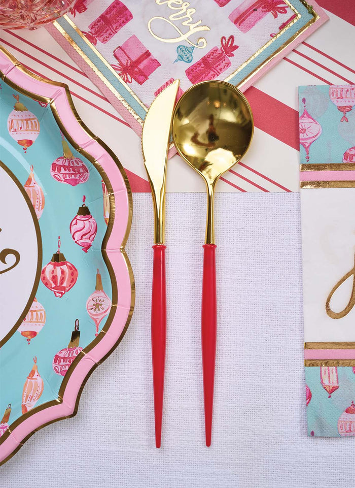 Sophistiplate's Red & Gold Bella Plastic Cutlery/24pc, Service for 8, features BPA-free gold knives and spoons with red handles, shown on a white cloth next to a decorative plate with pink ornaments and a festive blue napkin.