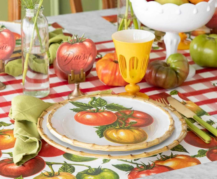 A table set with tomato-themed plates, a yellow glass, green napkin, gold cutlery, and a red and white checkered tablecloth. The Hester & Cook Red Painted Check Runner (20x25") plus decorative tomatoes with name cards add a festive touch.