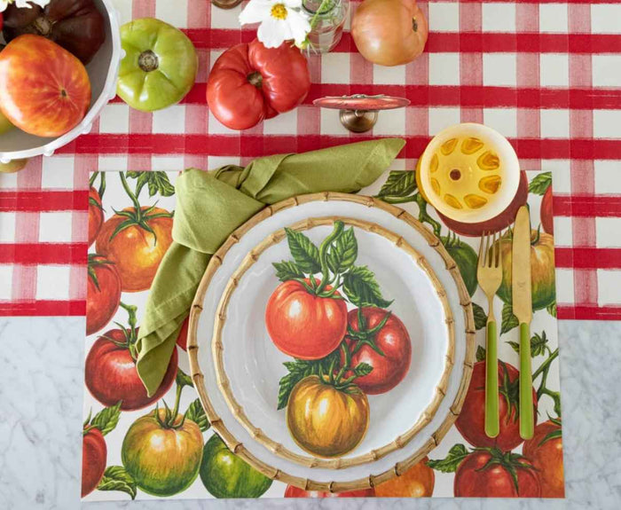 A place setting features a tomato-themed plate, gold utensils, green napkin with yellow ring on a colorful placemat and is set atop Hester & Cook’s Red Painted Check Runner (20x25"), accented by tomatoes, flowers, and a playful table runner.