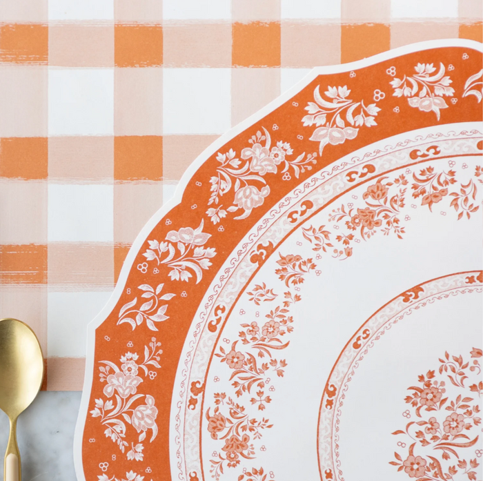 An ornate orange and white floral plate rests on a marble surface next to a gold spoon and fork, atop Hester & Cook’s Regal Pumpkin Placemat (12 Sheets), against an orange and white checkered tablecloth.