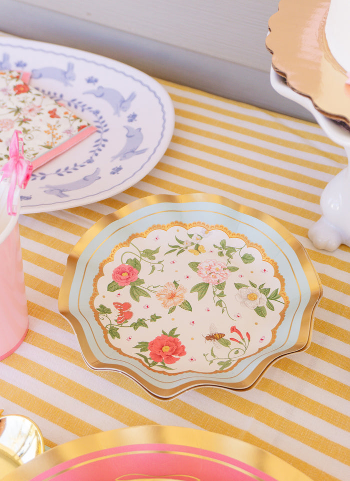 The Rococo Table Setting by Sophistiplate LLC features a yellow and white striped tablecloth, designer gold-rimmed plates, a white plate with a blue bunny, and matching floral napkins—perfect for a chic party-in-a-box.