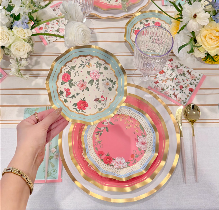 A hand holds a Rococo Appetizer/Dessert Bowl from Sophistiplate above an elegant tablescape with layered floral plates, gold cutlery, crystal glasses, and white and yellow flowers on a white tablecloth.