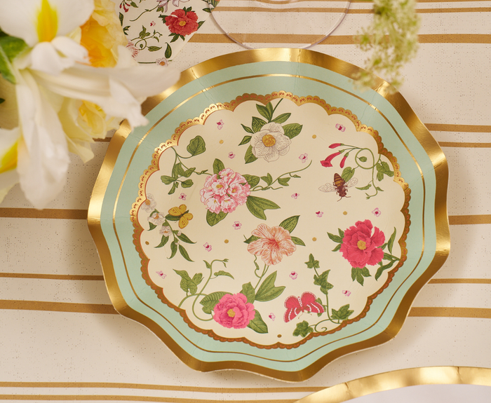 The Rococo Table Setting by Sophistiplate LLC features scalloped disposable plates with gold rims, adorned with pink, white, and yellow flowers, green leaves, and a bee—displayed on a cream tablecloth with gold stripes beside white and yellow blooms.