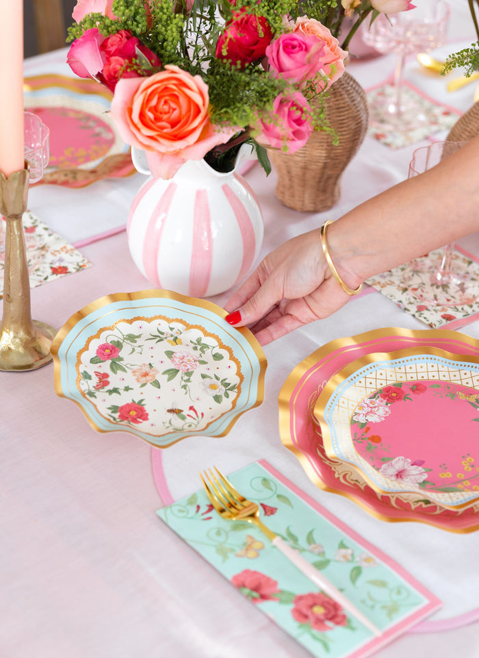 A hand sets a Rococo Table Setting by Sophistiplate LLC on a pink table with gold utensils, floral napkins, layered pink and gold plates, and a striped vase of colorful roses and greenery at the center.